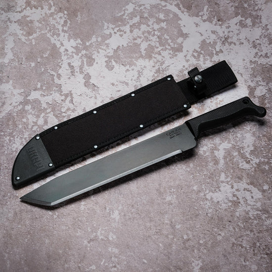 Okapi O-Tanto machete with canvas sheath resting on rugged grey stone floor
